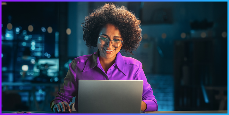 A women smiling as she sits working on a laptop.