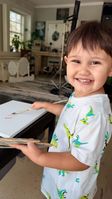 A smiling young boy wearing a dinosaur t-shirt holding a wooden painter's palette and touching a paintbrush with red paint to a blank white canvas.